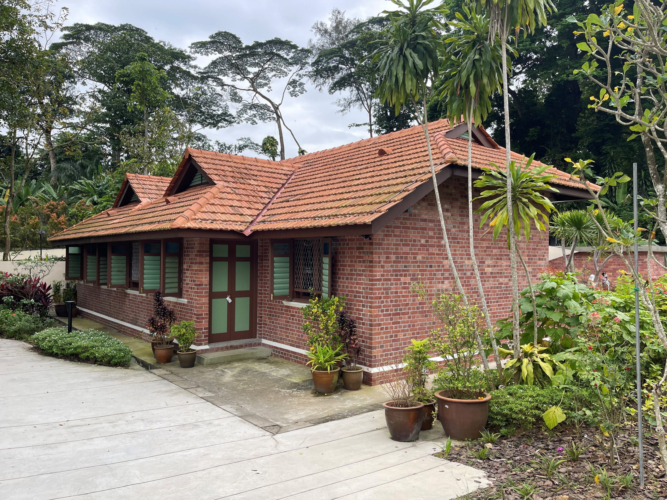 A brick house with a red-tiled roof surrounded by plants and trees on a cloudy day.